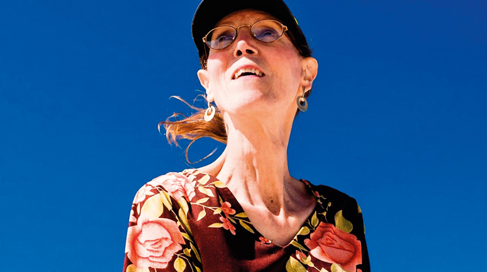 Foto mostra mulher olhando para cima com cabelo preso ventando. Céu azul de fundo