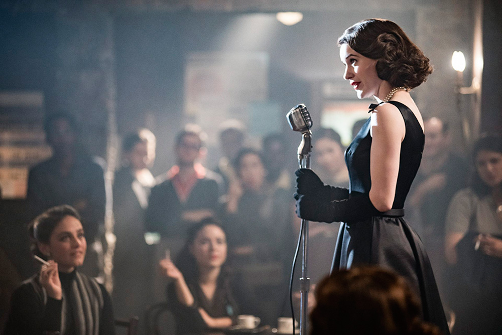 Uma mulher, no cenário e com roupas dos anos 1950, segura um microfone em frente a uma plateia