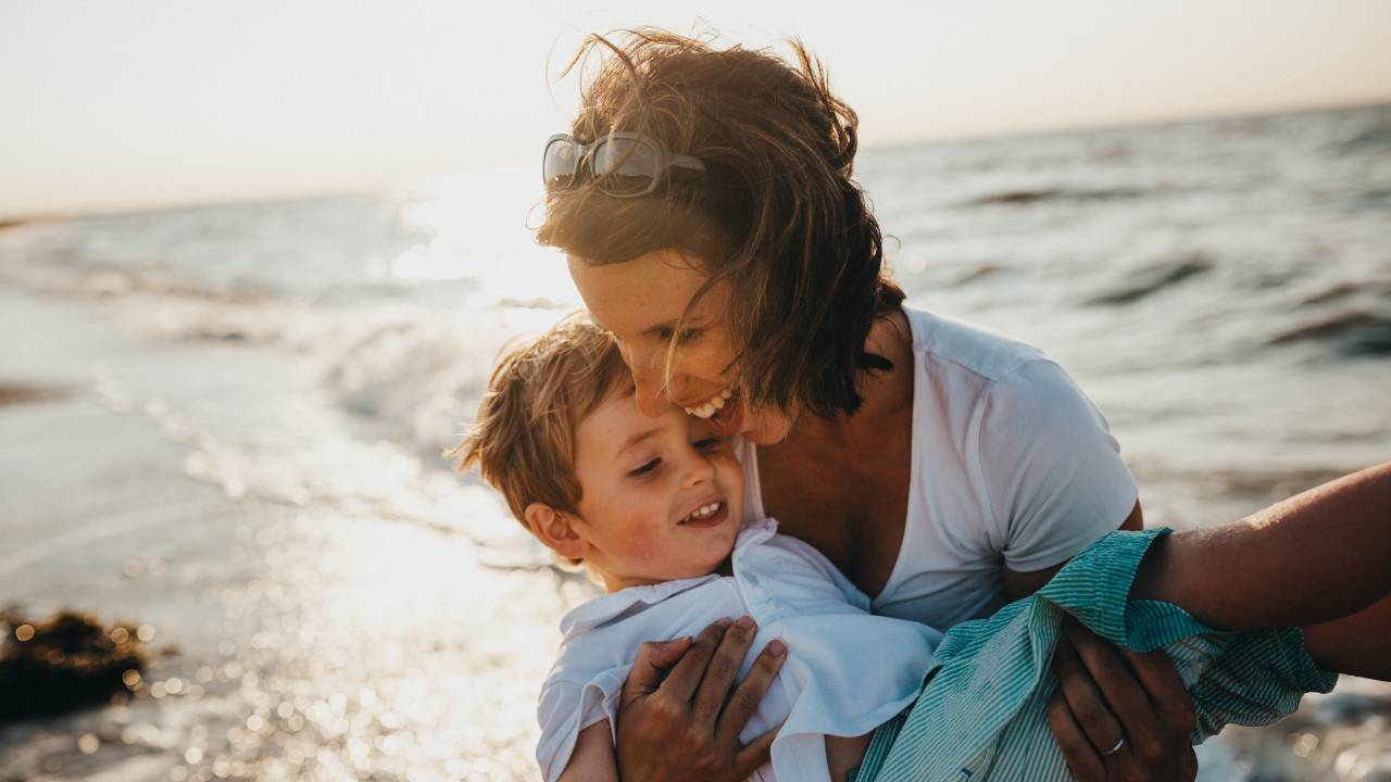 Mulher abraça criança em seu colo, ambos com sorriso no rosto, na praia