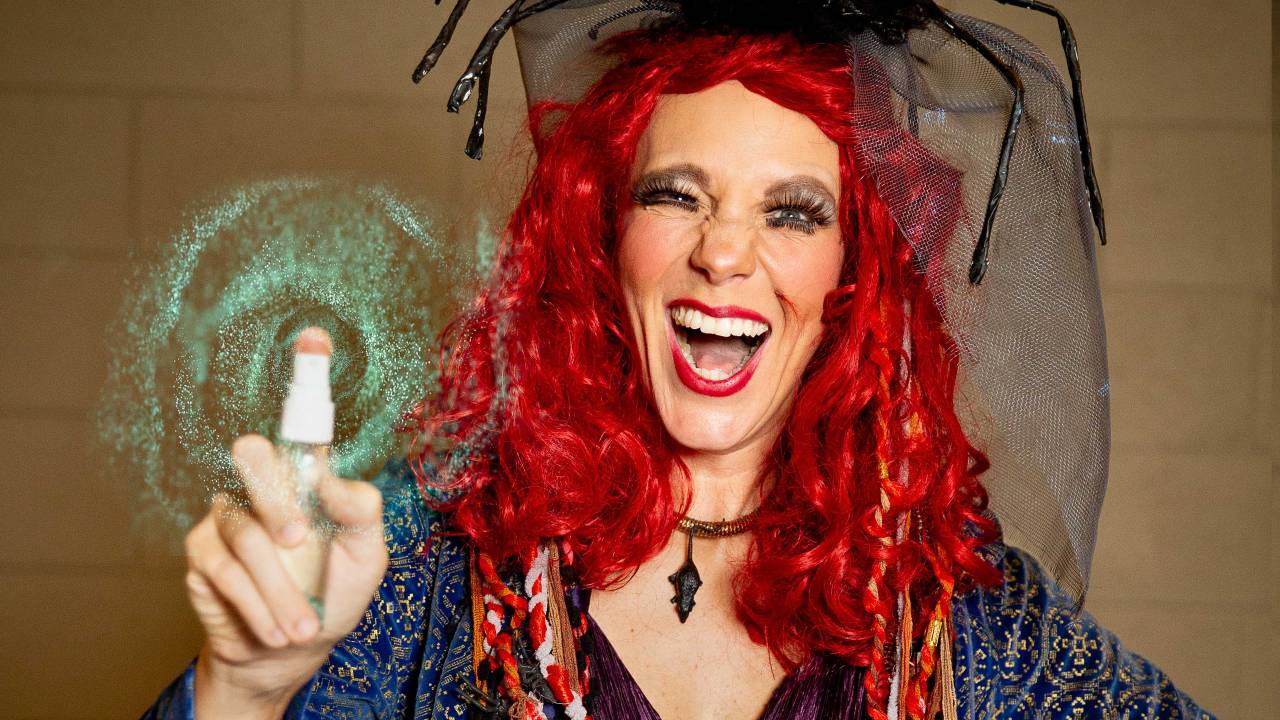 A atriz Carol Hubner, fantasiada de bruxa, com cabelos vermelhos e chapéu de aranha, pisca para a câmera, fazendo um "feitiço" com o dedo, de onde sai um efeito visual verde