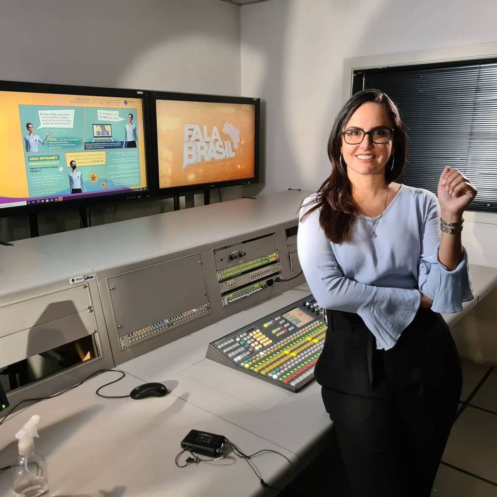 A foto mostra a jornalista Carla Cecato dentro de uma sala de operação de TV, sorrindo e posando para a câmera