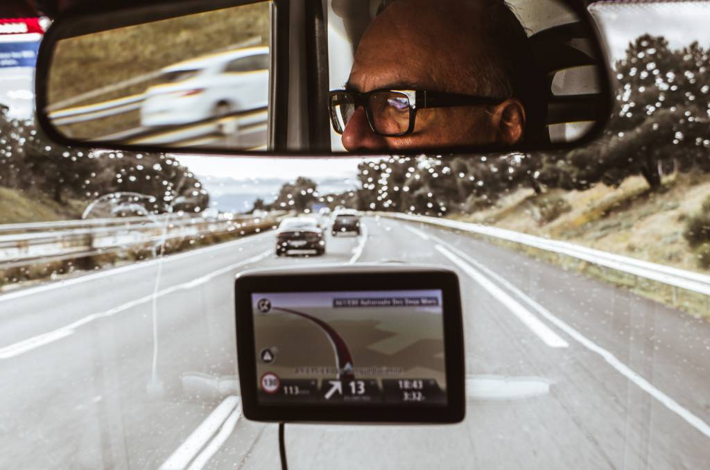 Rosto de Arnaldo Danemberg com óculos de grau aparecem no retrovisor de um caminhão. Em frente, a foto exibe a estrada e um GPS.