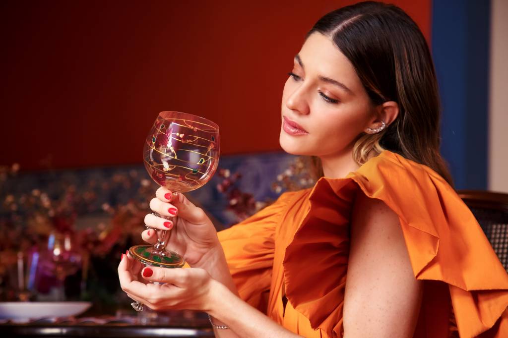 Atriz Luma Costa posa com vestido laranja e segura uma taça de vidro decorada com desenhos coloridos.
