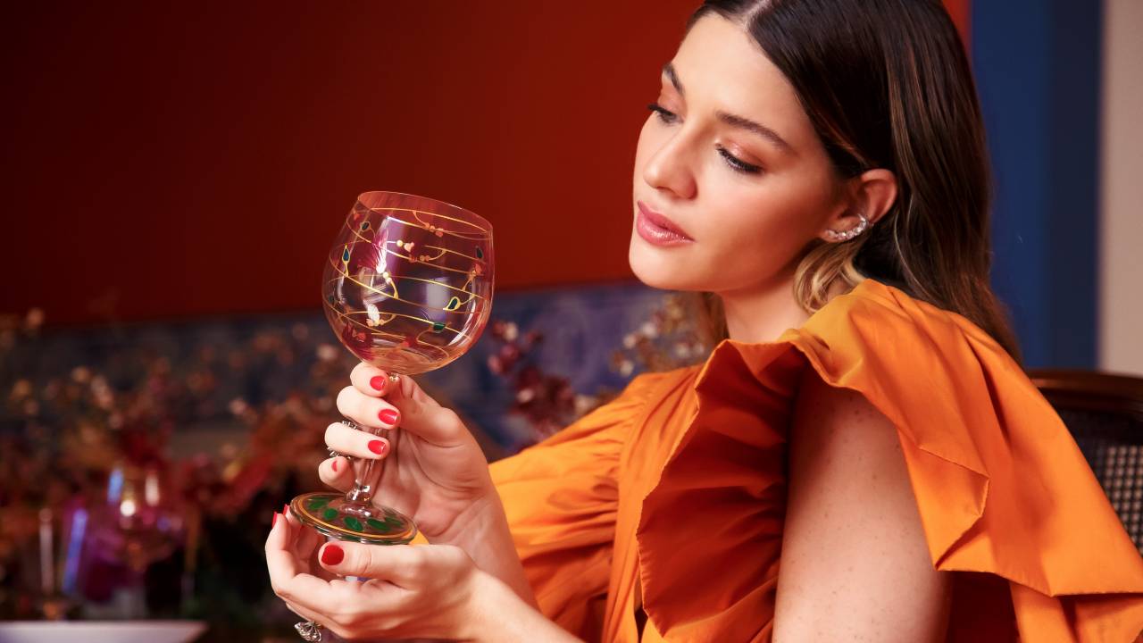 Atriz Luma Costa posa com vestido laranja e segura uma taça de vidro decorada com desenhos coloridos.