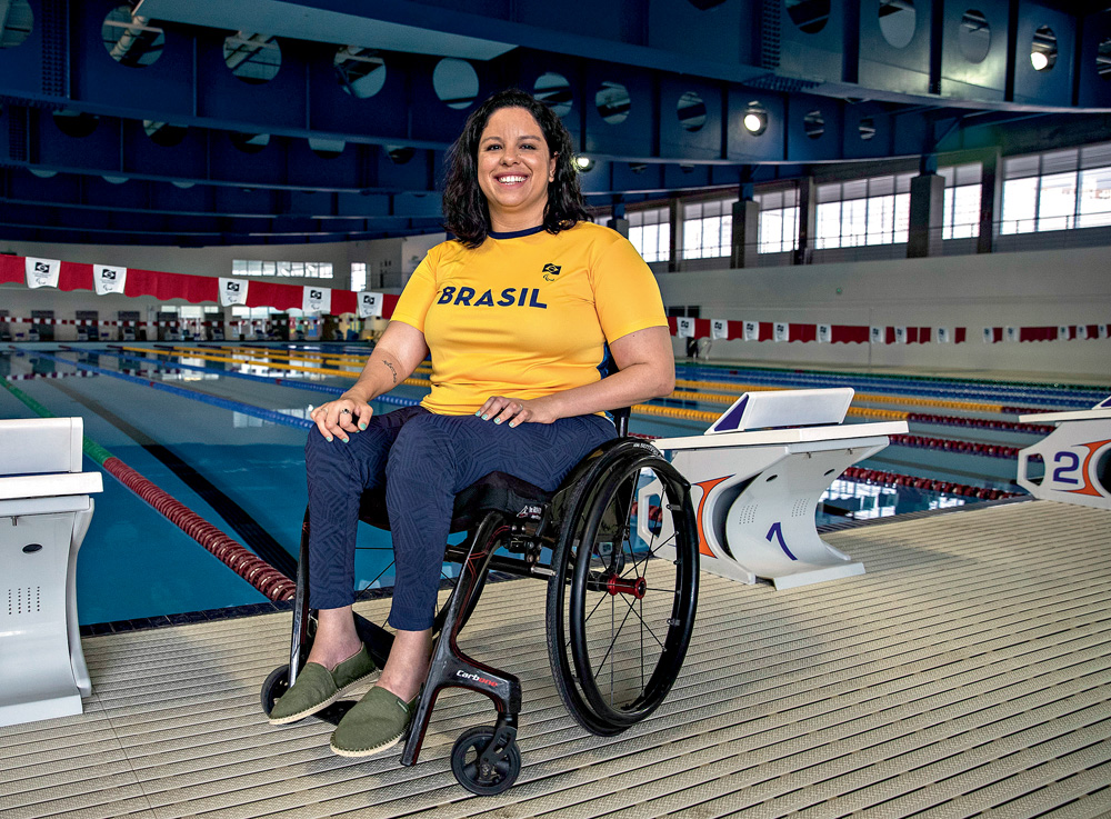 A fotografia mostra Edênia, de cadeira de rodas, em frente à uma piscina de natação. Ela está com a camisa amarela escrito Brasil e sorrindo para a foto.