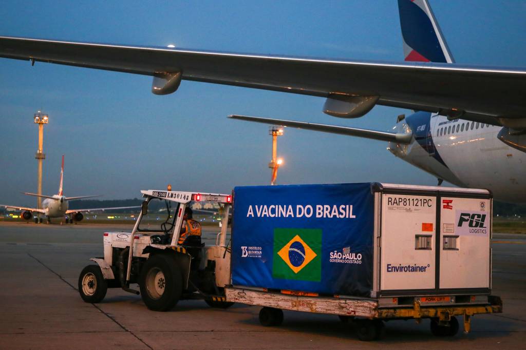 Imagem mostra trator transportando contêiner estampado com logo do Brasil, em que se lê "a vacina do Brasil", acompanhado de logotipo do Instituto Butantan e do governo de São Paulo. Trator e contêiner estão ao lado de avião