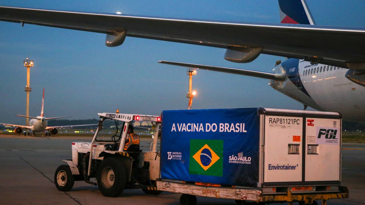 Imagem mostra trator transportando contêiner estampado com logo do Brasil, em que se lê "a vacina do Brasil", acompanhado de logotipo do Instituto Butantan e do governo de São Paulo. Trator e contêiner estão ao lado de avião