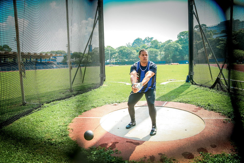 A imagem mostra o Allan em um campo de treinamento de arremesso de martelo. Ele está uniformizado e girando o martelo em um dia ensolarado.