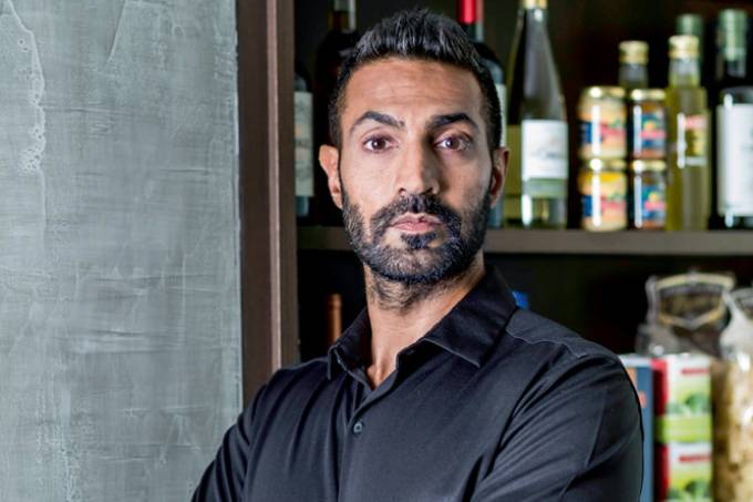 Alykhan Karim posa de braços cruzados com prateleira de vinhos ao fundo. Usa barba e uma camisa longa preta.