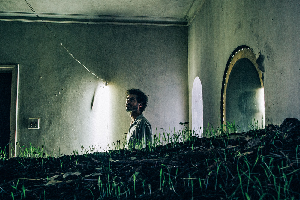 Homem olha para o teto de uma casa. Ao seu lado, uma estrutura horizontal com terra e plantas