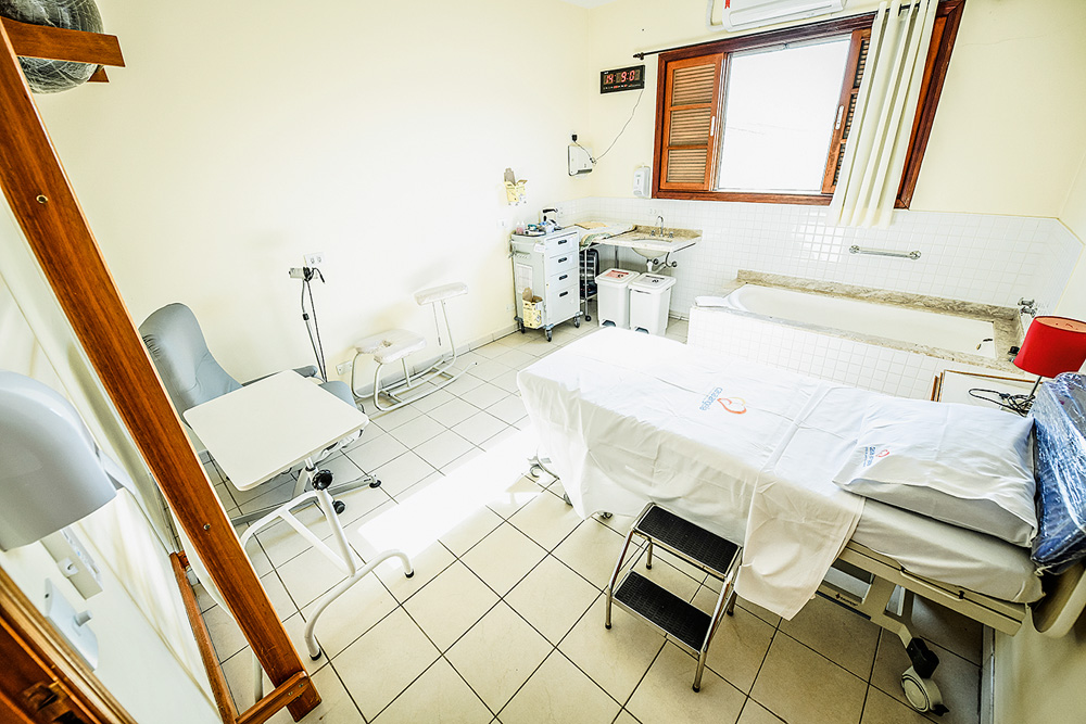 Sala de parto na Casa Angela. Maca, banco, pia e aparelhos médicos. Foto tem luz da claridade mostrada pela janela