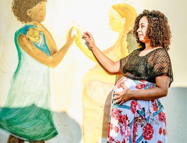 Mulher grávida coloca uma mão na barriga e a outra em uma pintura, em uma parede, de uma mãe com bebê no colo