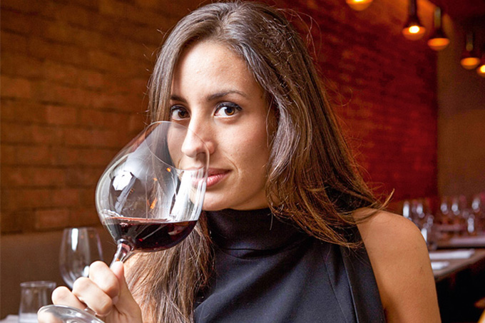 Mulher de cabelo castanho longo e blusa preta em destaque bebendo taça de vinho tinto.