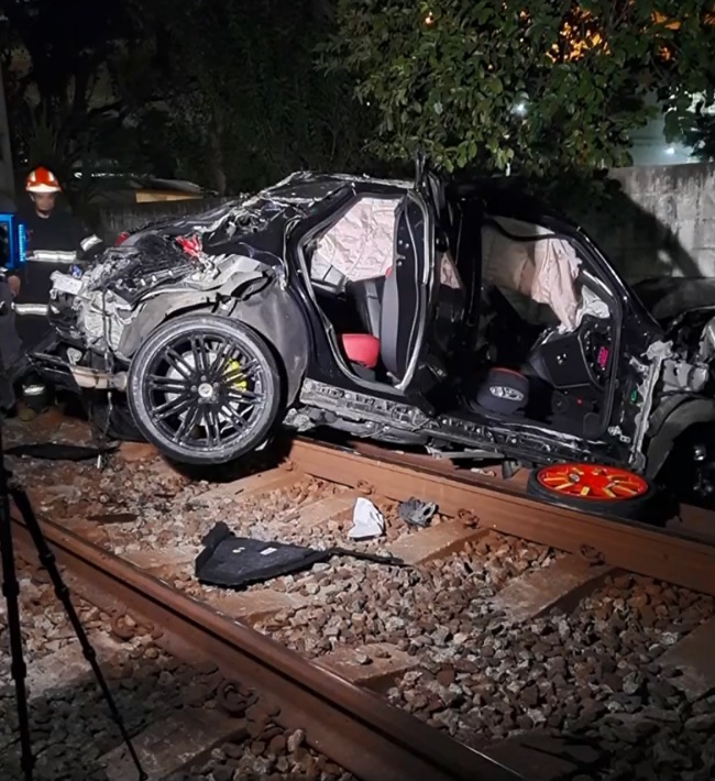 A imagem mostra um carro completamente desfigurado, inclinado para frente e com uma das rodas sobre o trilho de trem.