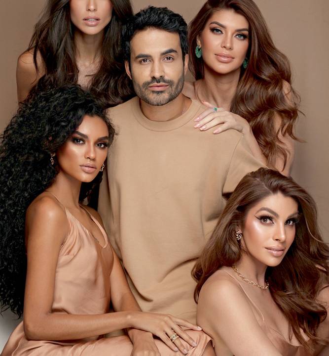 Cabeleireiro Chris Rodrigues posa rodeado de quatro modelos, todas com cabelos escuros. Todos da foto vestem roupas na cor bege, assim como o fundo do cenário.