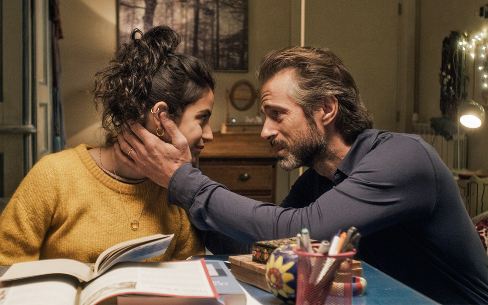 A imagem apresenta uma cena do filme italiano. Nela, um homem coloca a mão de maneira carinhosa no rosto de uma mulher enquanto ela sorri olhando nos olhos dele. Ambos estão sentados em volta de uma mesa.