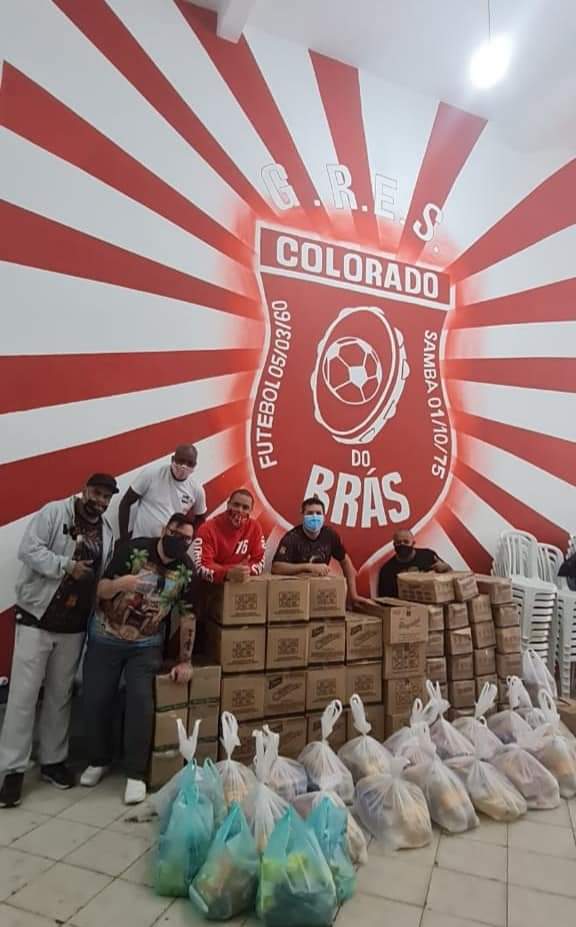 A imagem mostra seis pessoas de máscara com muitas caixas de papelão e sacolas em frente, de doações. No fundo, há uma parede com o escudo da escola de samba Colorado do Brás