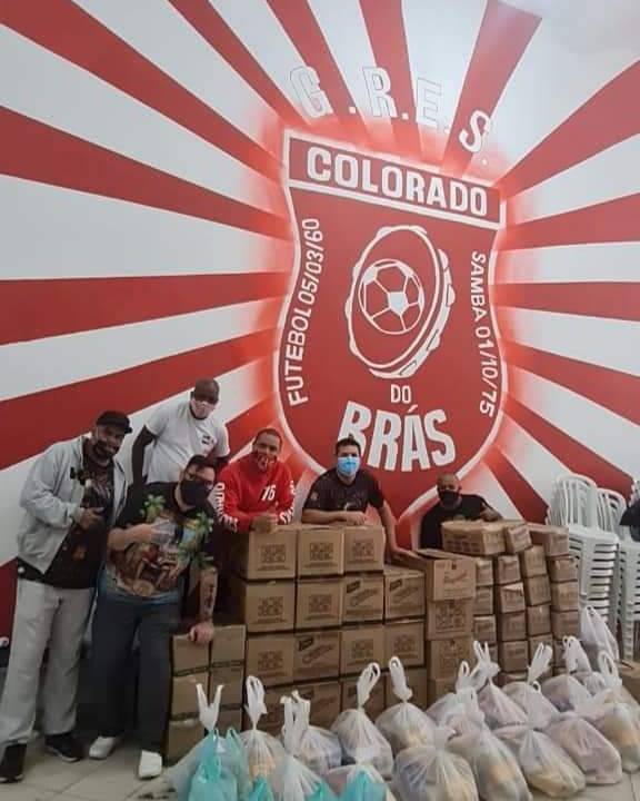 A imagem mostra seis pessoas de máscara com muitas caixas de papelão e sacolas em frente, de doações. No fundo, há uma parede com o escudo da escola de samba Colorado do Brás