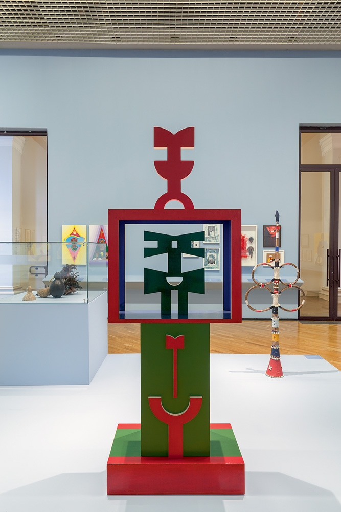 Escultura de Rubem Valentim em uma sala de museu. A obra é verde e vermelha e tem algumas formas geométricas