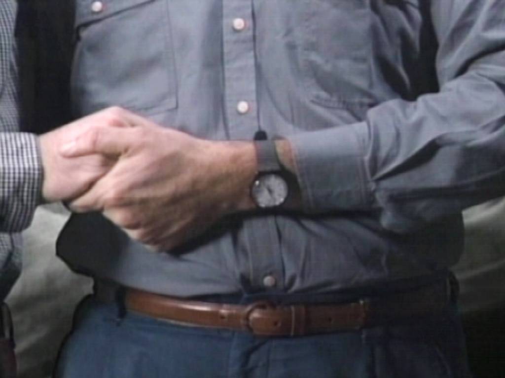 A imagem é uma foto na altura da barriga de um homem. No frame é possível ver a mão dele segurando a mão de outra pessoa com um relógio no seu pulso.
