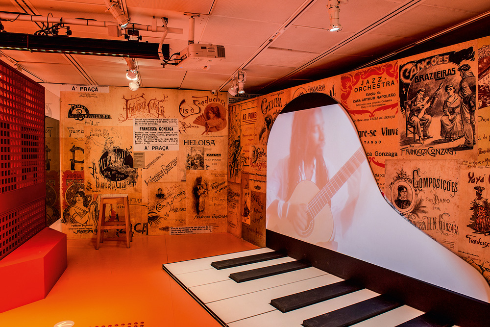 Uma sala em luzes alaranjadas exibe um telão, com formato de piano, em que um homem toca violão. Nas paredes, escritos e pinturas sobre música