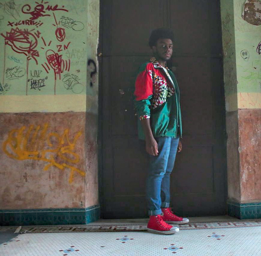 Jovem negro posa em frente a uma porta, em cenário com pinturas de tinta nas paredes