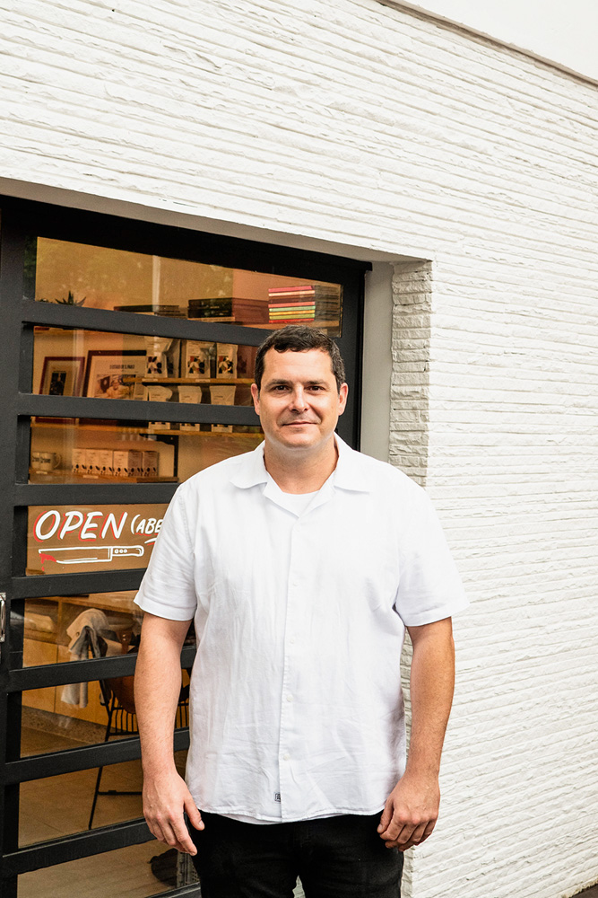 Homem ao centro (Rafael Protti) vestindo uma camisa branca larga com meio sorriso em frente à fachada da Crime Pastry Shop.