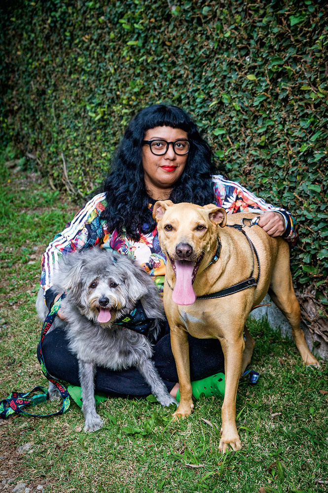 Uma mulher de cabelos bem pretos abraça dois cachorros, um cinza e um amarelo, na grama