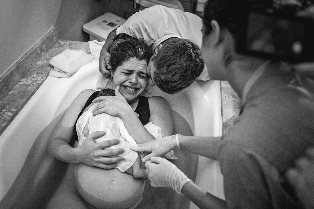 Foto em preto e branco mostra mulher na banheira dando a luz à bebê. Neném está em seu colo, um homem a abraça e ela chora