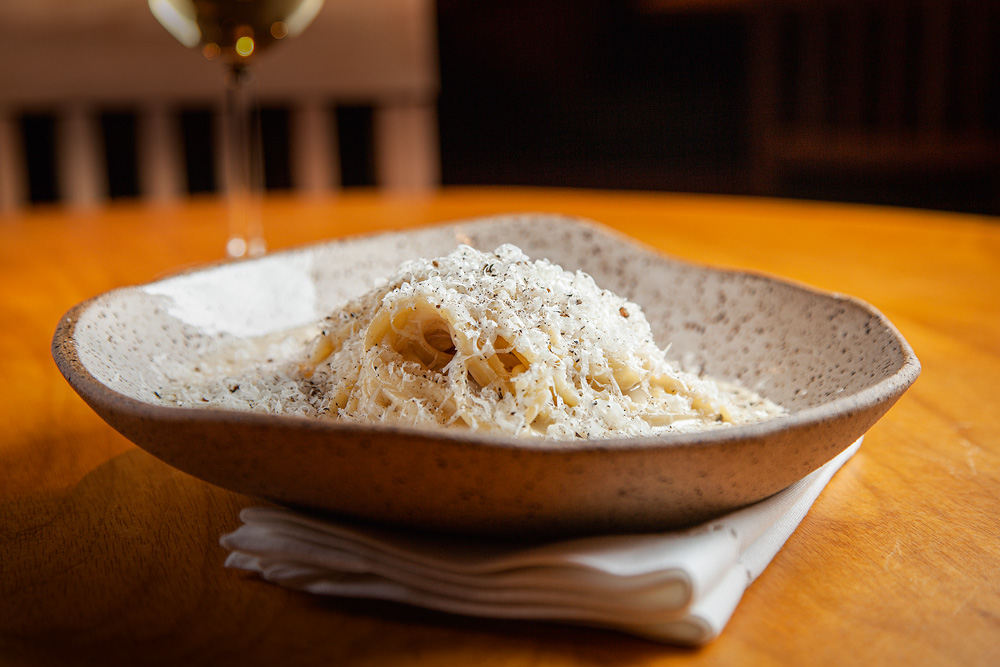 Espaguete cacio e pepe em um prato de cerâmica sobre guardanapo branco dobrado em cima de mesa de madeira. 