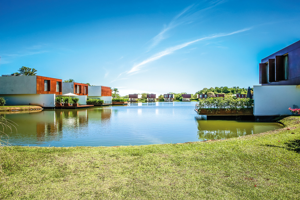 Imagem é um retrato amplo de uma área externa do condomínio de luxo. Há grama, um lago e grandes casas nas margens.