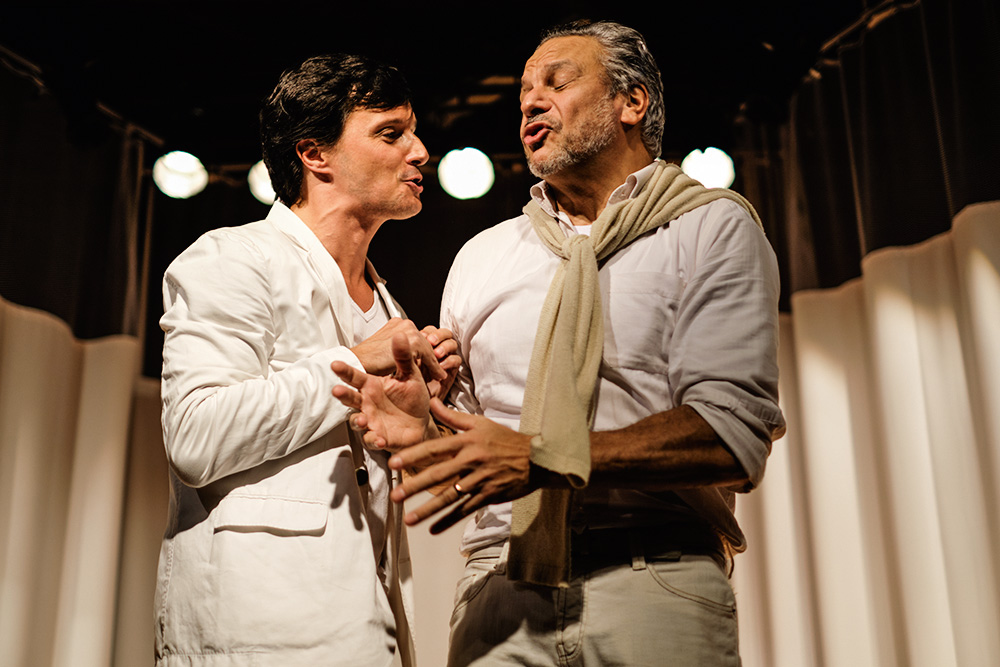 Dois homens, um grisalho e outro moreno, com roupas brancas, conversam no palco de um teatro