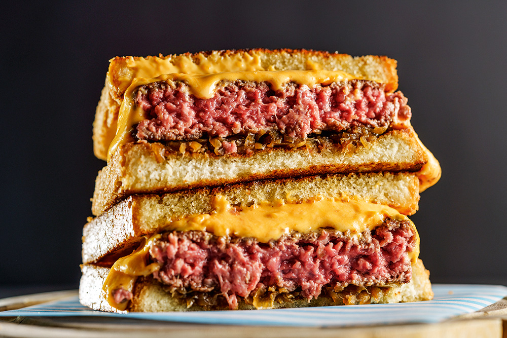 Foto do sanduíche "prensadão" do Fat Cow. Duas metades de hambúrguer no pão de forma, uma sobre a outra, com a carne rosada e bastante queijo cremoso aparente.
