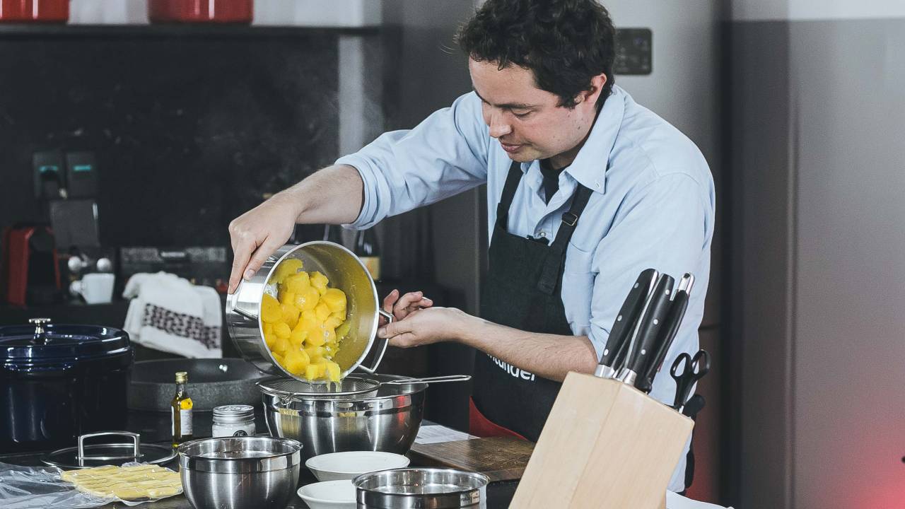 Chef Raphael Despirite em cozinha, atrás de bancada despejando uma panela de mandioquinha cozinha sobre escorredor de inox.