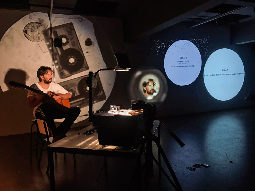 Homem toca violão sentado em uma cadeira. Atrás dele, projeções de fitas de música
