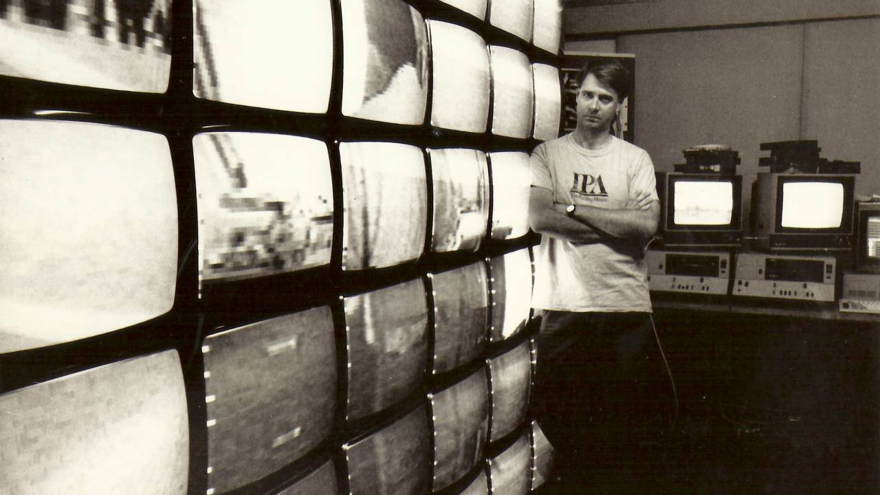 A imagem é antiga e mostra Rafael, de braços cruzados, apoiado em uma parede de televisores sorrindo para a câmera.