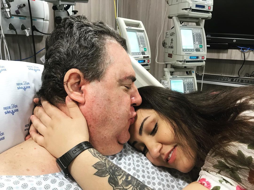 Na imagem é possível ver Gerson deitado em uma cama de hospital. Sua filha está deitada no seu peito enquanto ele beija a testa dela.