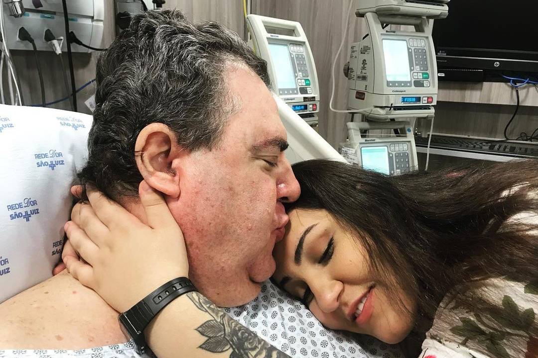 Na imagem é possível ver Gerson deitado em uma cama de hospital. Sua filha está deitada no seu peito enquanto ele beija a testa dela.