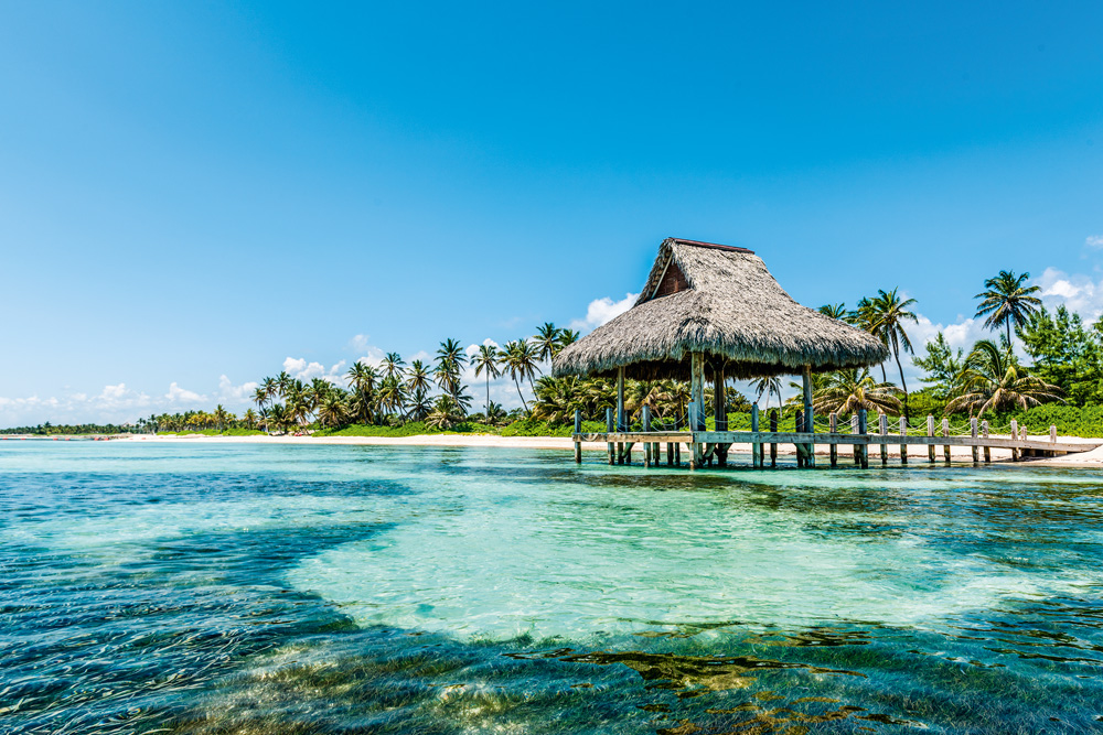 praia de punta cana, com uma faixa do mar aparecendo e a praia, com espécie de bangalô, ao fundo