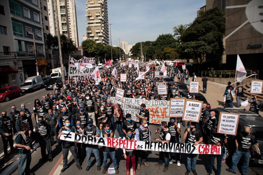A imagem apresenta uma rua cheia de pessoas segurando cartazes favoráveis ao protesto dos metroviários e criticando o governador Doria.