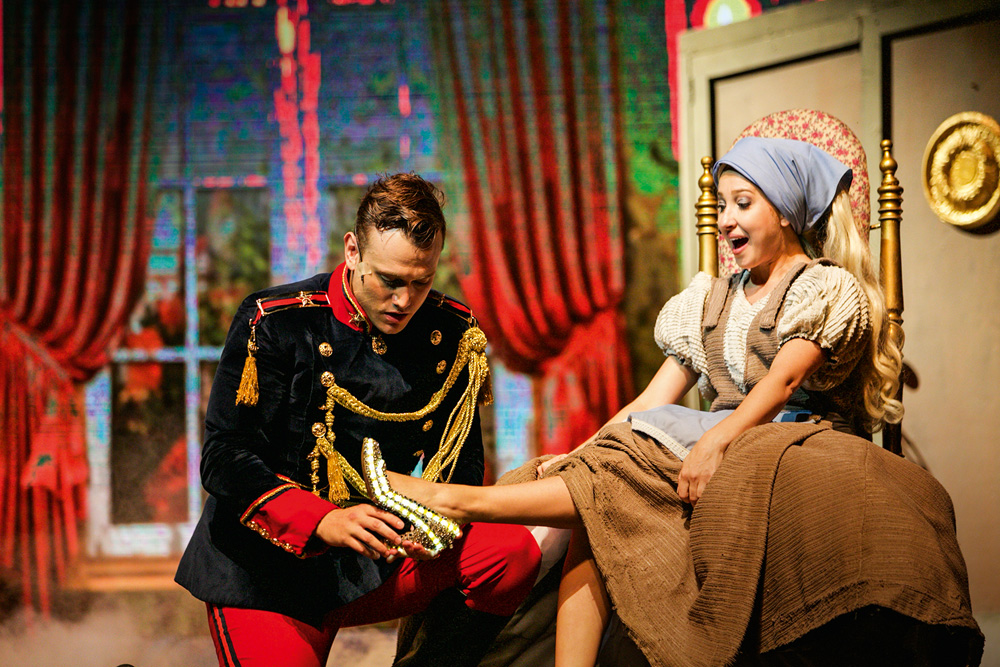 Mulher vestida de Cinderela, sentada num banco, prova sapato pelas mãos de um homem, o Príncipe. Eles estão ambientados em um palco de teatro
