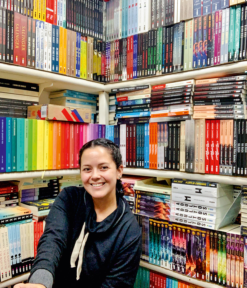 Heloiza Daou sentada posando para a foto junto de estantes repletas de livros ao fundo.