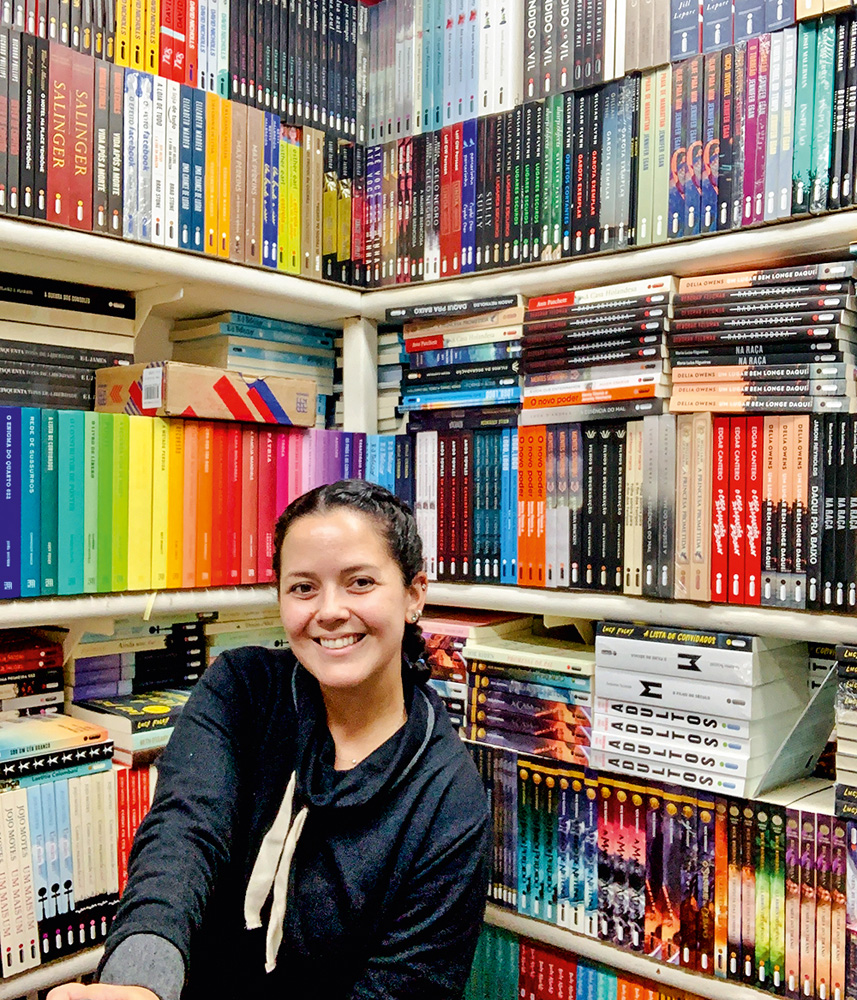 clubes-de-assinaturas-Heloiza-Daou-intrínseca.jpg Heloiza Daou sentada posando para a foto junto de estantes repletas de livros ao fundo.