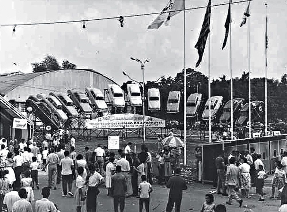 A imagem em preto e branco mostra a entrada do salão do automóvel. É uma fachada lotada de pessoas e, ao fundo, há carros erguidos com a frente voltada para o chão.