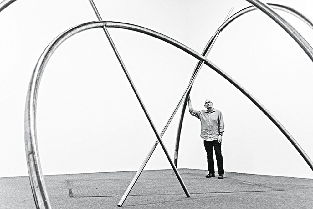 Foto mostra o artista José Resende ao lado de uma de suas obras. Há hastes em metal com fios enovelados nas pontas e vão do chão até outro ponto do chão arredondadas