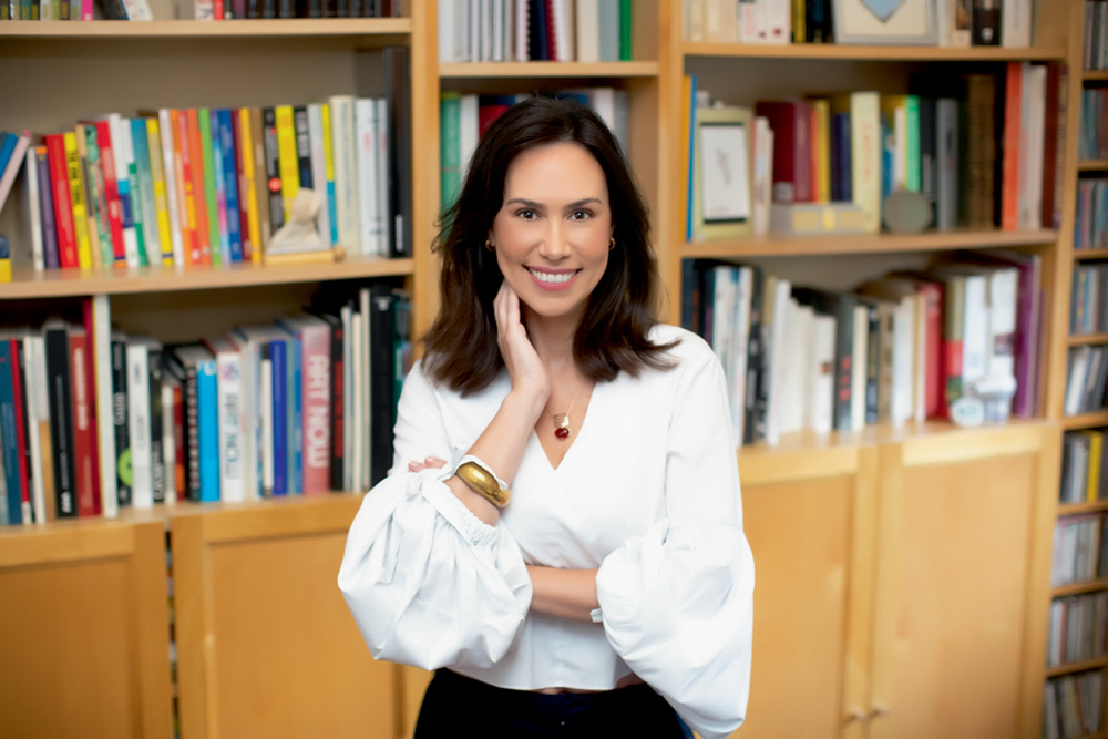 Mulher (Vivian Wolff) posa em frente a uma estante de livros. Sorrindo