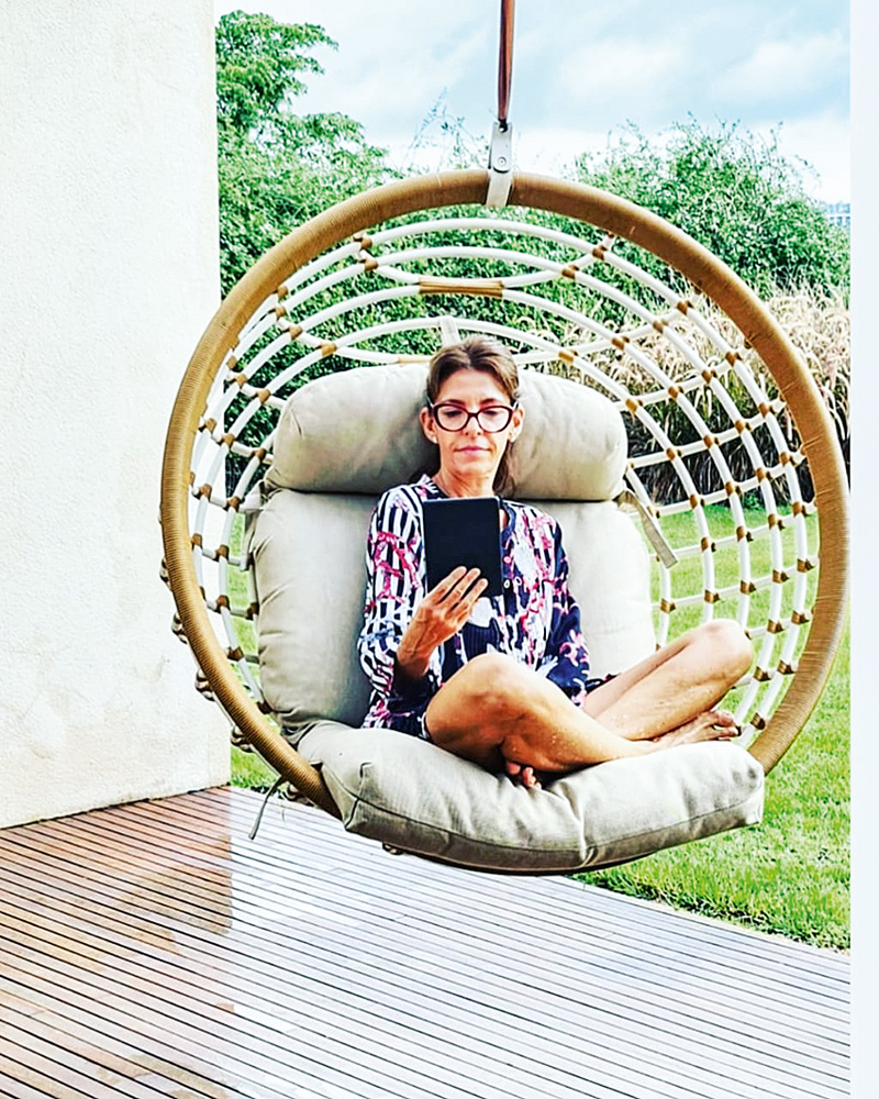 A imagem mostra Ligia Danesi sentada em um sofá exótico perto da área externa da sua casa enquanto mexe em um tablet com a mão esquerda.