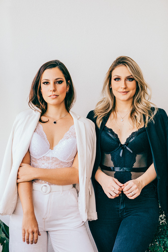 Karol e Karina, da LingeBox, posam para a foto de pé. Karol veste peças brancas e Karina peças pretas, ambas com peças de lingerie inclusas no look.