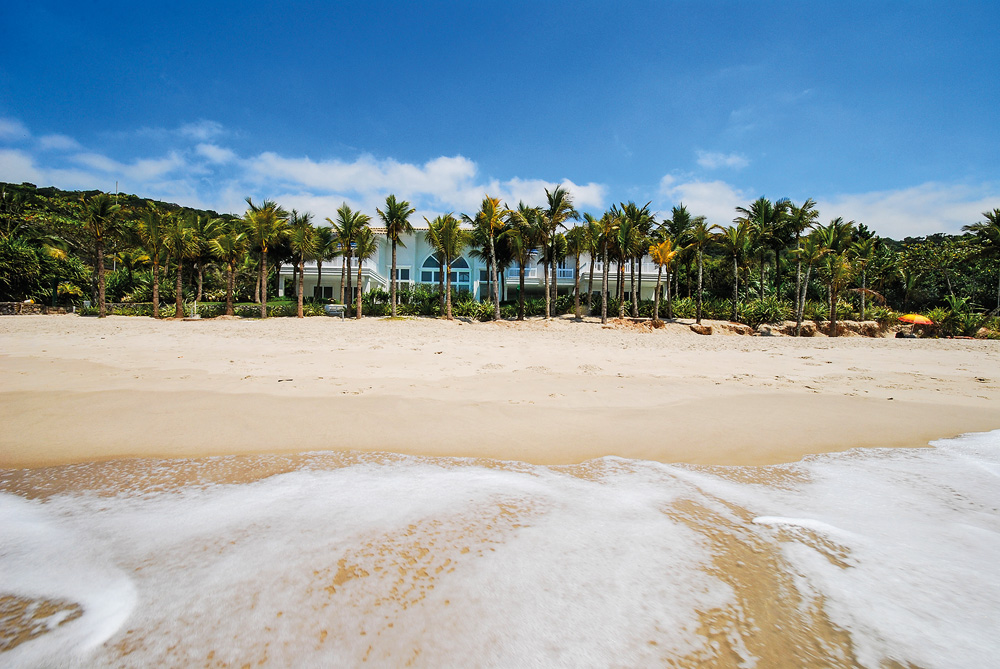 A imagem é vista a partir do mar, na praia. Nela, dá para ver no horizonte entre várias árvores uma mansão bem na margem da areia.