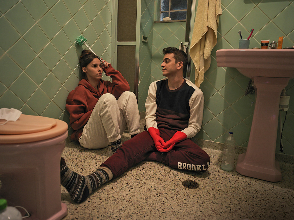 A imagem mostra Victor Lamoglia e Thati Lopes sentados no chão de um banheiro, olhando um para o outro em cena do filme